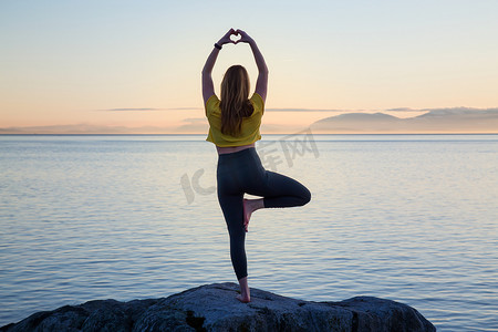 年轻女子在充满活力的日落期间在一个岩石岛上练习瑜伽。