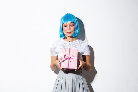 可爱生日背景摄影照片_戴着蓝色派对假发的可爱生日女孩的形象，看着礼品盒，站在白色背景上