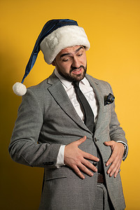 吃得太多的年轻英俊的大胡子男人戴着蓝色的圣诞老人帽子和西装摸着肚子，看着黄色背景中孤立的相机。