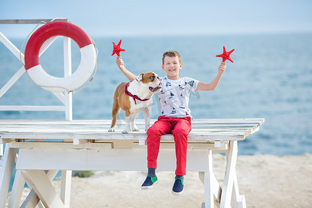 英俊的男孩青少年快乐地与他的朋友斗牛犬在海边度过时光孩子狗抱着玩两个海星靠近救生圈漂浮穿着红色裤子裤子拖鞋和 T 恤