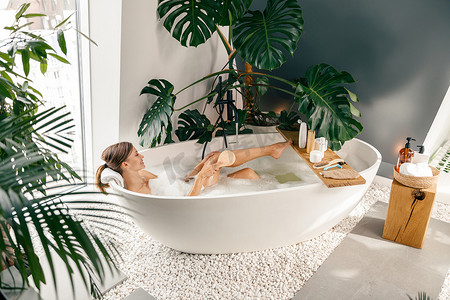 在用热带植物装饰的现代浴室里泡泡浴时，放松的年轻女性用刷子
