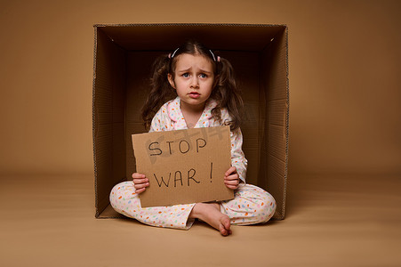 停止战争摄影照片_心烦意乱的小女孩坐在一个纸箱里，展示一张停止战争的海报。