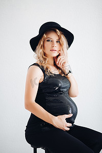 戴着黑色帽子和闪闪发光的上衣的时尚孕妇抱着她的肚子。