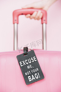 粉红色背景下，人手拿着带行李标签的旅行手提箱