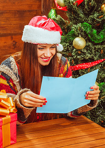 身穿休闲温暖毛衣、头戴圣诞帽的漂亮白人妇女在圣诞树旁读圣诞贺卡，上面有几个闪闪发光的球，她喜欢读圣诞祝福。