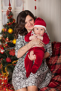 穿着圣诞老人服装的妈妈和儿子在靠近一棵圣诞树的家庭环境中靠着一堵白色的木墙。