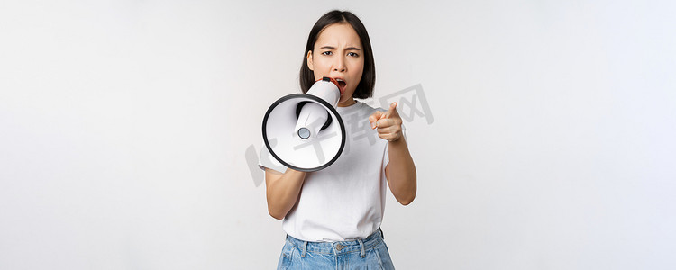 愤怒的亚洲女人拿着扩音器，骂人，指责某人，用扩音器抗议，站在白色背景上