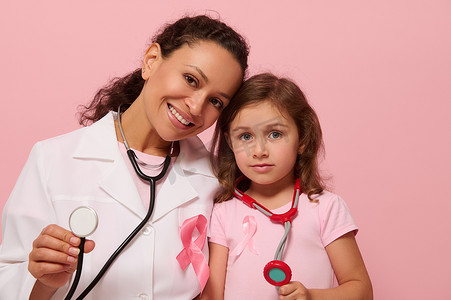 国际卫生组织摄影照片_微笑的混血医生和小女孩，脖子上都挂着乳腺癌意识粉红丝带和电话内窥镜，向镜头展示听诊器，隔离在粉红色背景中，复制空间。