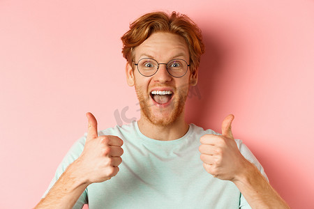 戴着眼镜和 T 恤的快乐红发男人的脸，竖起大拇指，看起来很兴奋，赞同和赞美酷炫的促销活动，站在粉红色背景上