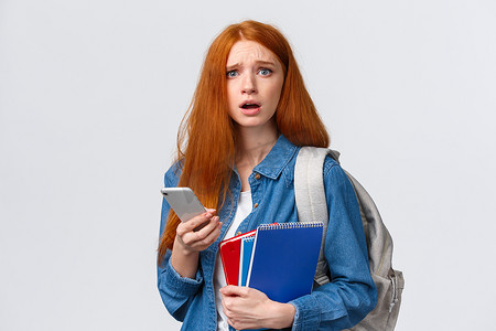 不安而悲伤的阴郁可爱的红头发女性看起来陷入困境的相机，在智能手机显示屏上阅读令人担忧的东西，拿着手机、背包和笔记本，就像在大学里一样