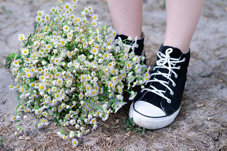 鞋子夏季摄影照片_夏季，在自然背景下，运动鞋附近无法辨认的女性腿和户外的白色野花