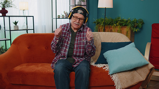 戴着无线耳机的喜出望外的老人在家里客厅舒适的沙发上跳舞、唱歌