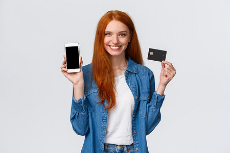 腰上肖像自信的女性，长着红色长发，蓝眼睛，拿着信用卡并展示智能手机显示屏，轻松的银行服务，支付账单互联网，微笑满意