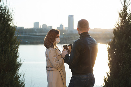 年轻夫妇，一男一女戴着医用口罩和手套，在街上用一次性杯子喝咖啡，在河流和城市的背景下互相看着对方。