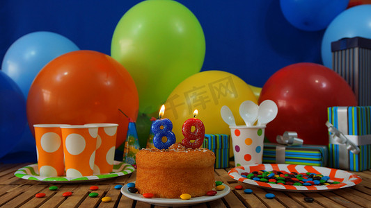 气球墙摄影照片_质朴木桌上的生日蛋糕，背景是彩色气球、礼物、塑料杯和塑料板，背景是糖果和蓝墙
