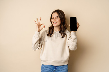 微笑的快乐女孩展示智能手机屏幕应用程序、移动界面、好的标志，站在米色背景上