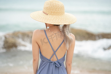 身穿蓝色连衣裙、头戴草帽的女人的背面，站在海边的一块木头上。