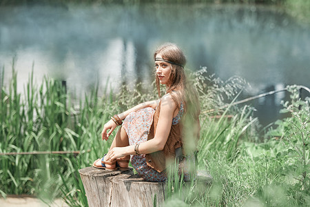 美丽的嬉皮女孩坐在树桩上看着池塘