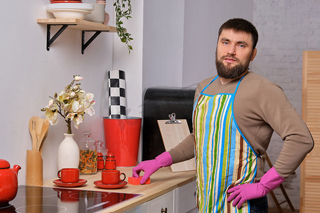 戴着围裙的男人摄影照片_厨房里留着胡子的年轻帅哥，戴着围裙和粉色手套，用洗涤剂清洁烹饪表面