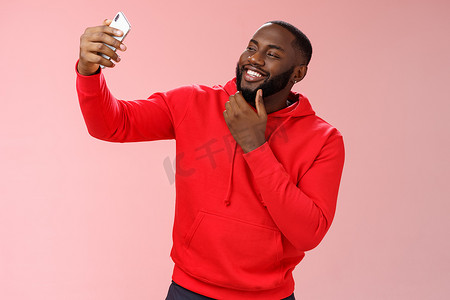 穿着红色连帽衫的厚颜无耻、酷酷的非洲裔美国人自信、留着胡须的男人拿着智能手机自拍更新日期应用程序个人资料照片，举起触摸脸，时髦的微笑看起来电话显示调情