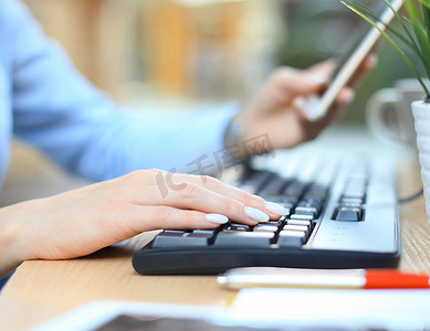 特写女性手在办公室的办公桌上使用智能手机在键盘上打字。