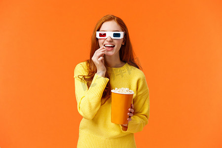 穿着黄色毛衣、面带微笑、古怪的红发女人，戴着 3D 眼镜在电影院看电影，吃着爆米花，用娱乐和兴奋的表情好奇地盯着看
