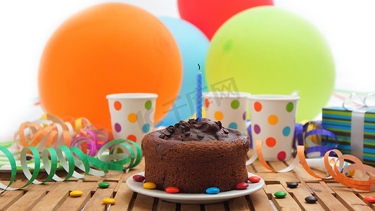 巧克力生日蛋糕，蓝色蜡烛熄灭在质朴的木桌上，背景是彩色气球、礼物、塑料杯、糖果和背景白墙
