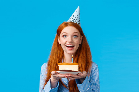 可爱生日蛋糕摄影照片_腰上肖像开朗乐观可爱的微笑红发女性女孩我生日帽，拿着一块生日蛋糕，兴奋地庆祝自己的生日，站在蓝色背景上的睡衣