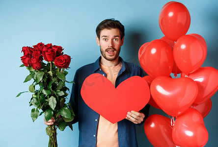 恋爱中的男人在浪漫约会时带来惊喜礼物，手捧红玫瑰和情人节红心花束，站在气球旁，看着情人，蓝色背景