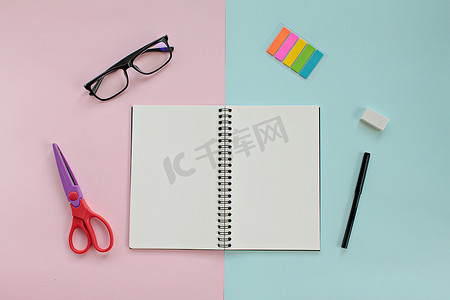 工作台的顶视图，配有空白笔记本、眼镜和办公文具，背景为粉色和蓝色，创意平躺模型设计
