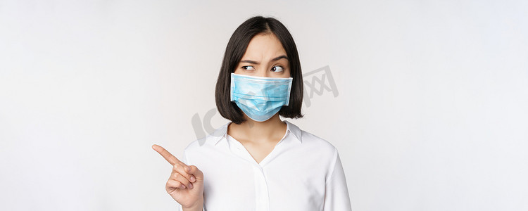 戴着医用面罩的亚洲年轻女性站在白色背景上，用可疑的困惑表情向左指着