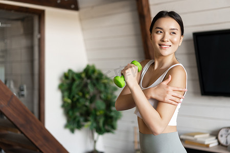 活跃健康的亚洲女孩，身体健康，在家做健身运动，举起哑铃，摸二头肌，在客厅锻炼