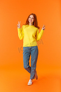 穿着牛仔裤和冬日黄色毛衣的垂直全长可爱红发少女站在橙色背景上，带着和平标志，微笑着摆出无忧无虑的快乐情绪