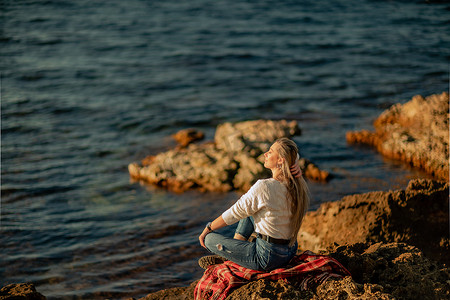 白毯子摄影照片_迷人的金发白种女人在日落时分在海滩上享受时光，坐在毯子里，看着一边，背景是落日的天空和大海。