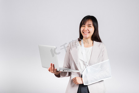 受伤女孩摄影照片_亚洲女商人在事故后自信地微笑着断臂，并戴上手臂夹板进行治疗，但仍然必须工作，她拿着笔记本电脑