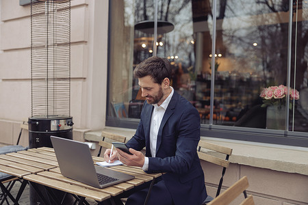 微笑的商人在咖啡馆的笔记本电脑上工作，拿着电话，在笔记本上记笔记。