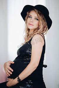 戴着黑色帽子和闪闪发光的上衣的时尚孕妇抱着她的肚子。