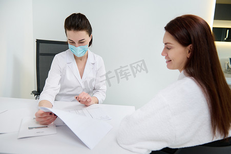 自信的戴医用口罩的女医生会诊她的病人，在白色办公室分析她的医疗记录。