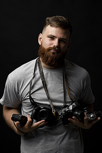 留着胡须的专业摄影师的肖像，身穿灰色 T 恤，手上和肩上拿着一堆不同的相机，看着相机，准备好拍摄。