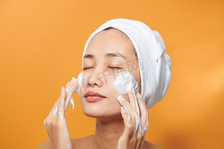 天然药摄影照片_温柔的半裸女人裹着毛巾，用橙色背景中隔离的泡沫清洁剂洗脸