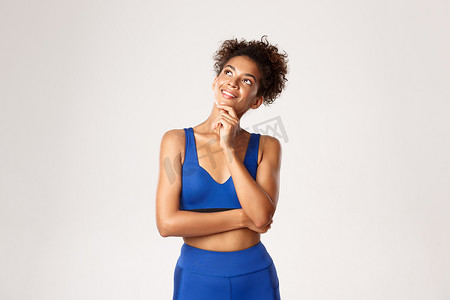 蓝色运动服摄影照片_身着蓝色运动服、面带微笑地看着左上角、考虑锻炼、白色背景的体贴迷人的非洲裔美国健身女性的工作室照片