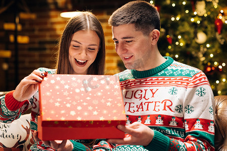 开朗的年轻女子打开圣诞礼品盒的肖像，脸上兴奋而惊讶，在除夕夜坐在家里靠近发光的圣诞树时感到高兴。