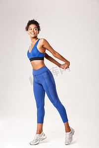 穿着蓝色运动服、微笑着的非洲裔美国女运动员在锻炼前伸展身体，向右看，站在白色背景上