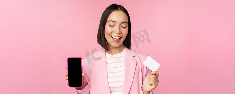 界面购物摄影照片_穿着西装、微笑的韩国女商人展示手机屏幕、信用卡、展示网上银行应用程序界面、粉红色背景