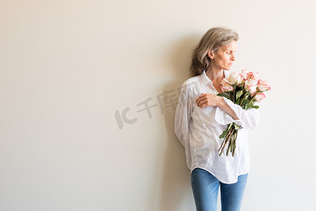 玫瑰花粥摄影照片_中年妇女的半长视图，头发灰白，白衬衫拿着粉色和奶油色的玫瑰
