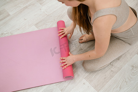 运动健身垫摄影照片_一位年轻女子在锻炼之前或之后滚动粉色健身垫或瑜伽垫，在家里的客厅或瑜伽室锻炼。