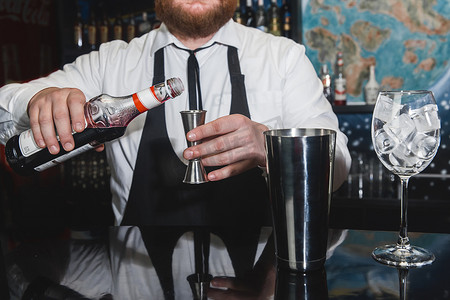 制酒摄影照片_专业调酒师的手将红色糖浆倒入量杯中，旁边是一个金属工具，用于准备和搅拌摇酒饮料和带有冰块的玻璃杯的酒精鸡尾酒