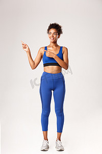 穿着蓝色运动服的全长非洲裔美国运动女孩，手指向左，看起来很高兴，推荐健身器材，站在白色背景上