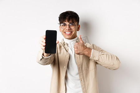 戴眼镜的英俊年轻人的肖像，显示空的智能手机屏幕和拇指，推荐在线应用程序或商店，高兴地站在白色背景上