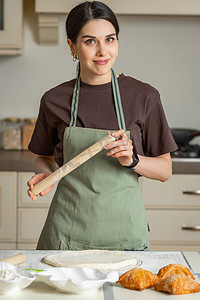 擀面杖摄影照片_美丽的黑发年轻微笑混血女人手里拿着擀面杖，穿着厨房围裙正在做饭